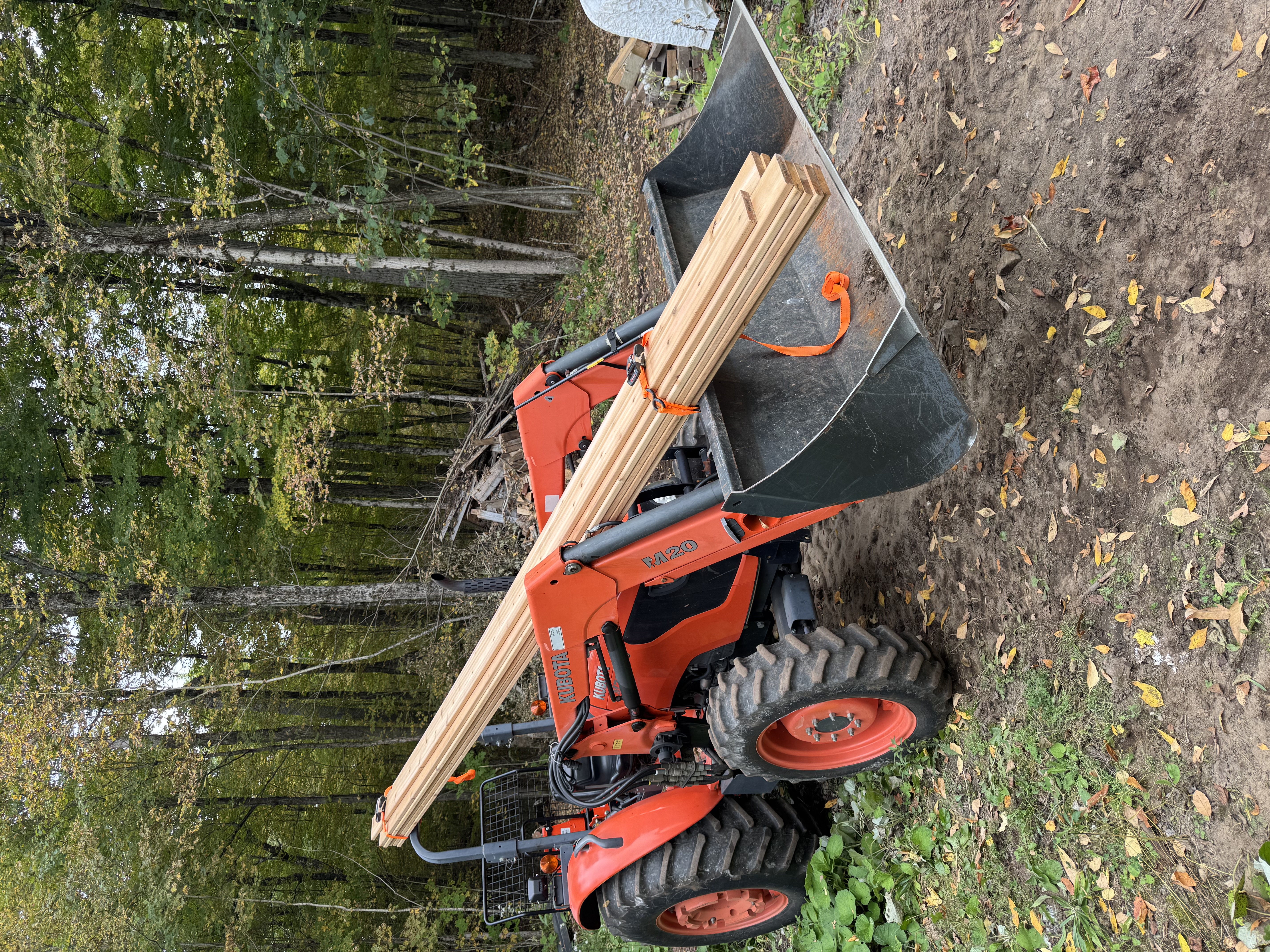 The tractor carrying a few 16 foot long lumber boards in an unusual manner: strapped to the bucket and hanging over the operator and strapped to the roll over bar, at the back of the tractor