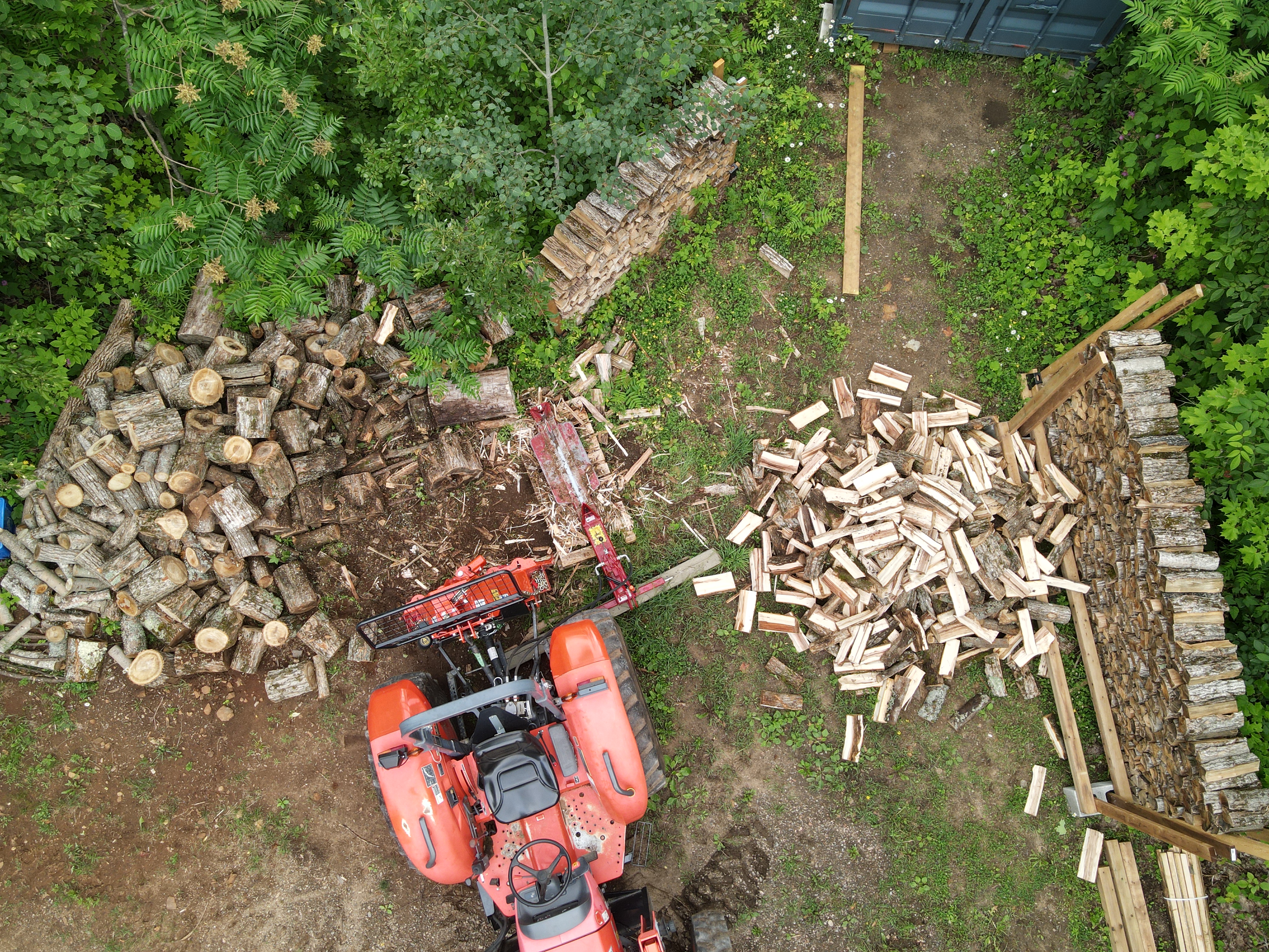 A drone view of splitting firewood, logs on the left and split firewood on the right. The splitter is not attached to the tractor the normal way but instead sitting beside it.