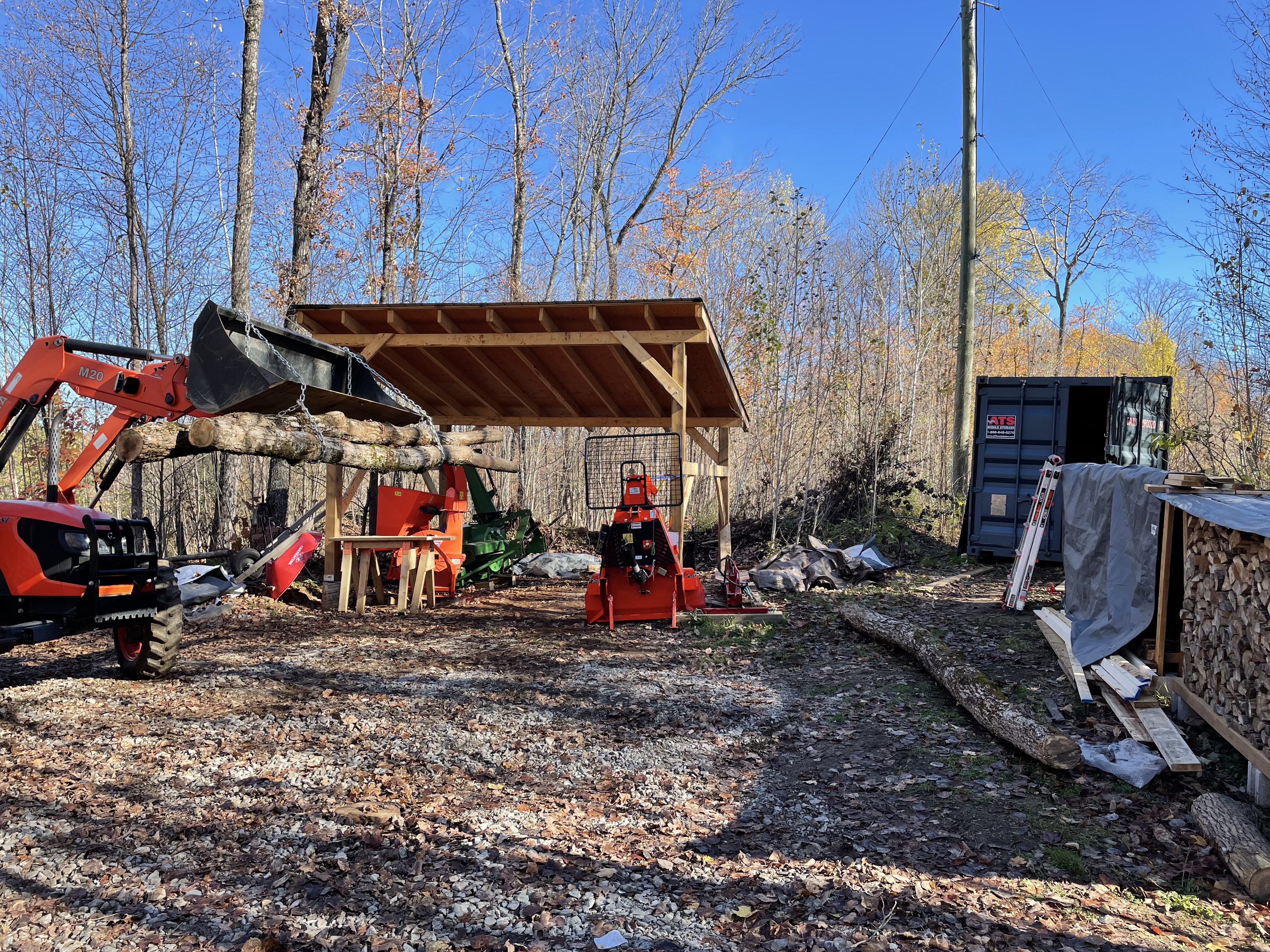 Tractor lifting up a bundle of logs with the bucket, using two chains wrapped around the logs. The tractor shed is in the background.