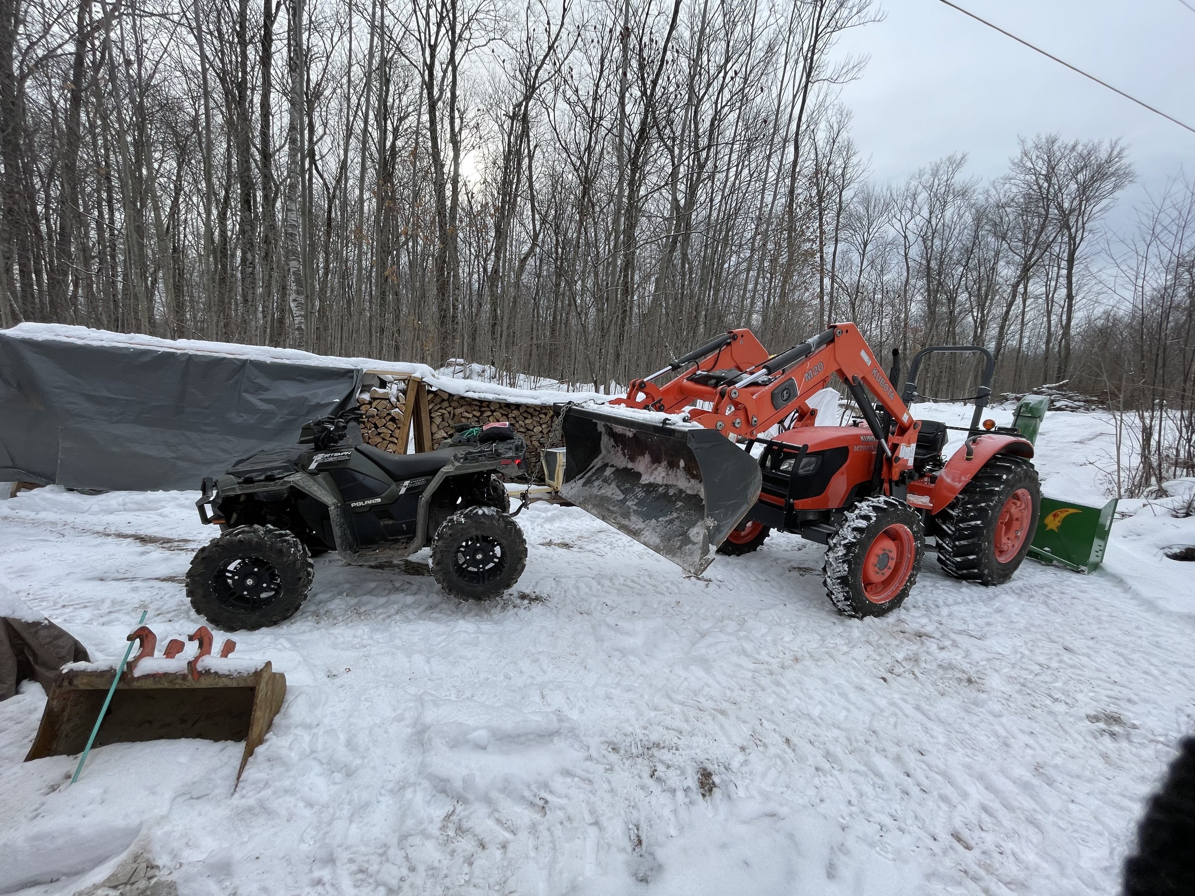 The ATV being lifted up by its rear end using the tractor loader and chains, on a snowy driveway, to install snow chains on the wheels.