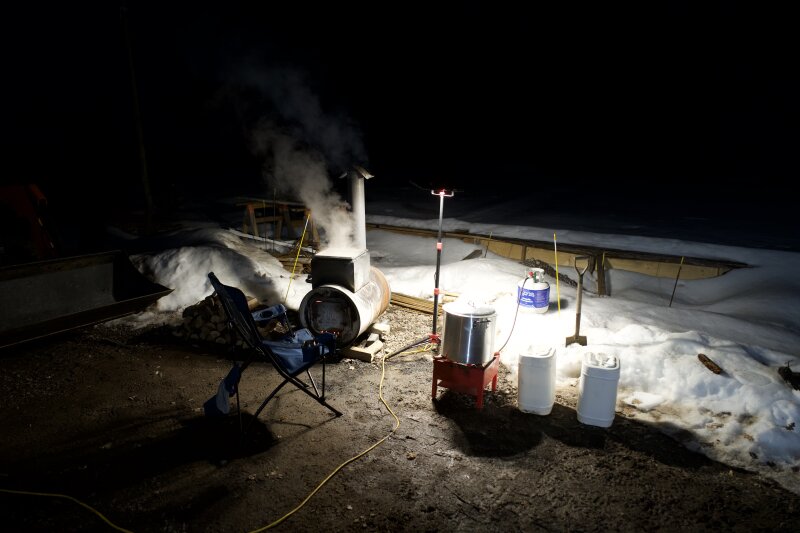 A dark evening with the barrel evaporator and finishing stove under a work light in the driveway. A camp chair sits in front of the mess.