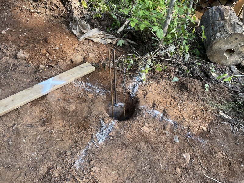 A shallow hole in the earth with visible bedrock a few inches down. Three pieces of rebar are epoxied into three drilled holes in the rock.