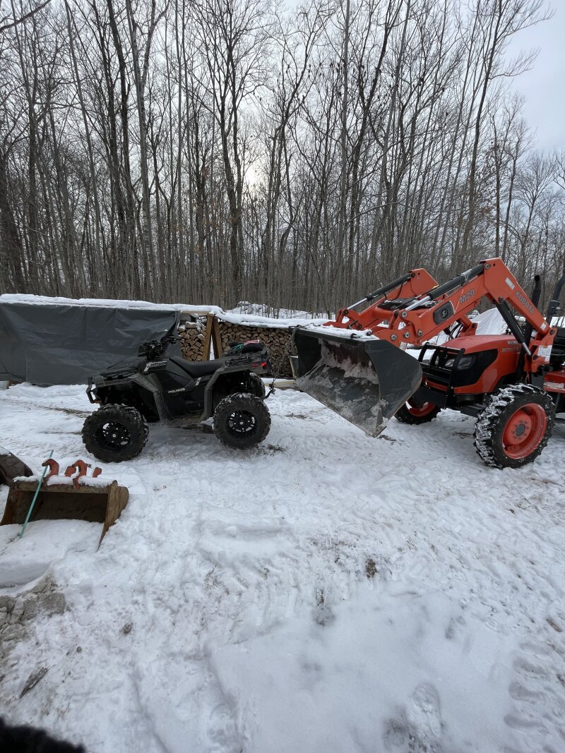 The tractor using its bucket to lift the rear of the ATV off the snowy driveway, allowing access to its tires to install snow chains