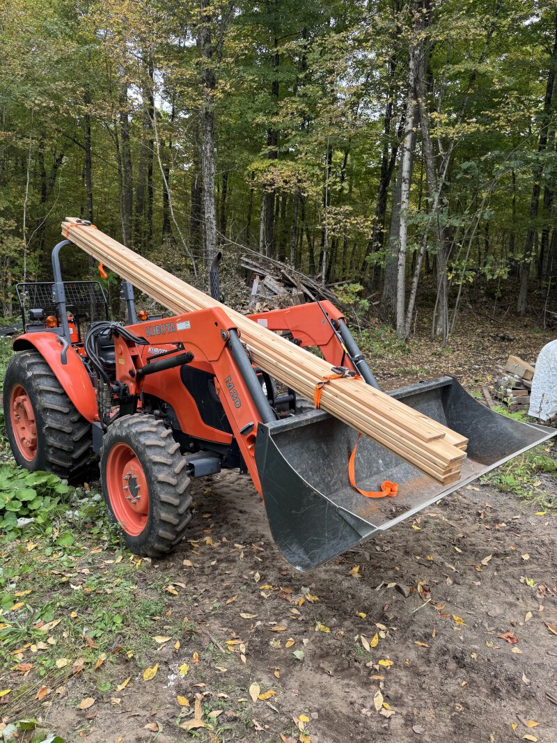 The tractor carrying a few 16 foot long lumber boards in an unusual manner: strapped to the bucket and hanging over the operator and strapped to the roll over bar, at the back of the tractor