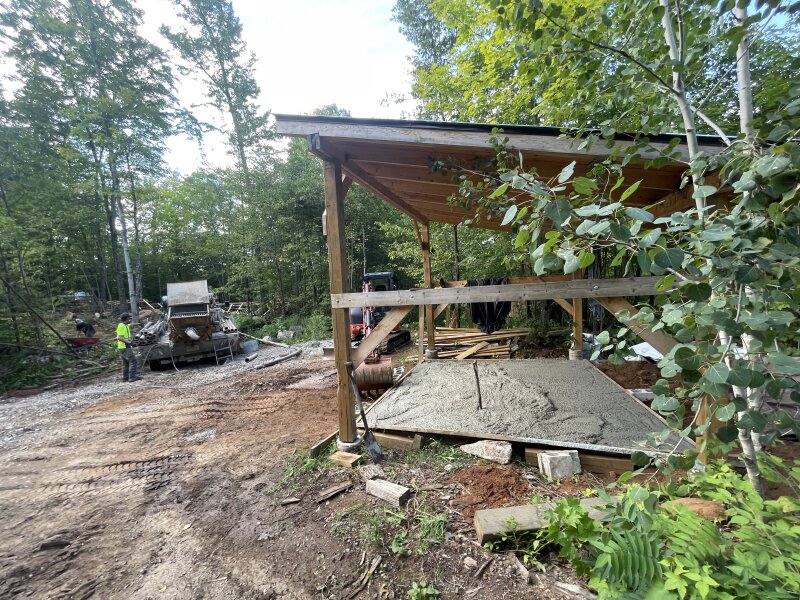 Concrete going into the hastily constructed forms under the shed roof, a concrete pumper and excavator visible in the background