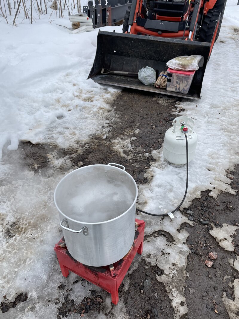A large propane burner with an even larger aluminum pot on it, in front of the tractor, on the snowy driveway.