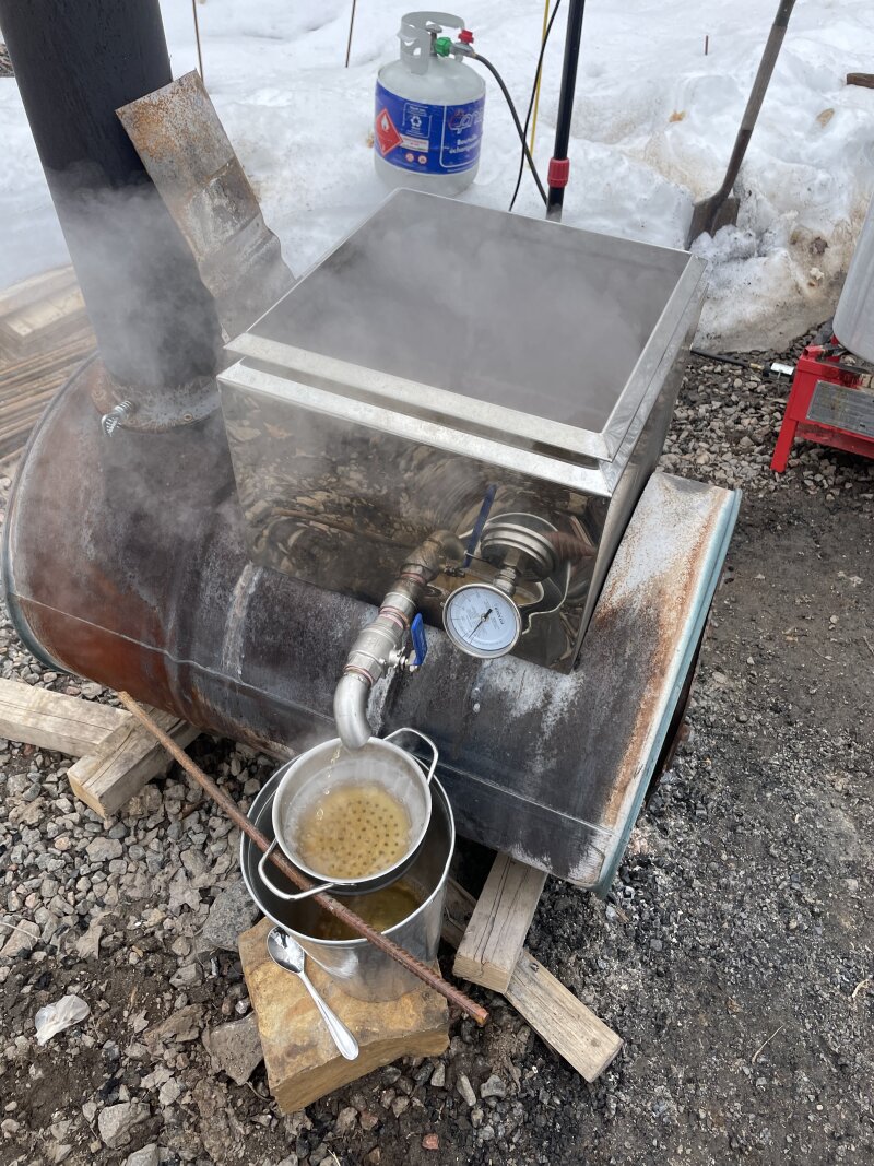 The draw-off valve on the evaporator pan slowly draining boiling partially-finished syrup into a rudimentary filter, cheese cloth in a strainer.